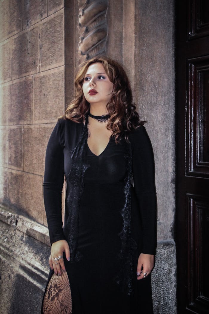 Woman in gothic attire standing against stone wall, conveying elegance and mystery.