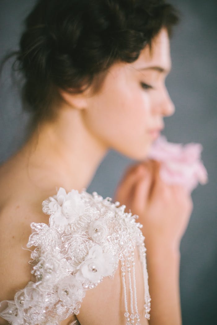 Soft-focus portrait of an elegant bride holding a pink flower, wearing embroidered lace.